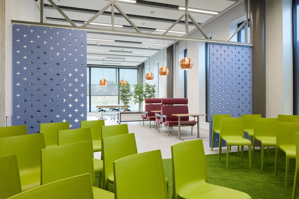 Doorkijkje vanuit een ruimte met groene stoelen en blauwe panelen naar een ruimte met bankjes en koperkleurige lampen.