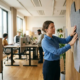 Kantoormedewerker installeert grijze akoestische panelen op moderne kantoorwand terwijl collega's werken aan zit-sta bureaus