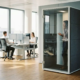 Glazen telefooncel met akoestische panelen in moderne kantooromgeving, medewerker belt terwijl collega's werken aan ergonomische bureaus