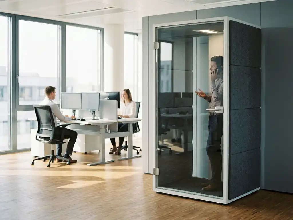 Glazen telefooncel met akoestische panelen in moderne kantooromgeving, medewerker belt terwijl collega's werken aan ergonomische bureaus