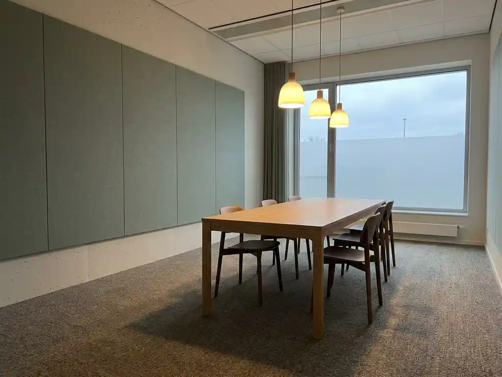 Lege moderne vergaderruimte met lichthouten tafel, grijsgroene akoestische wandpanelen en zachte hanglampen in natuurlijk daglicht.