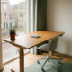 Zit-sta bureau met eikenhouten blad en ergonomische stoel bij raam met uitzicht op Nederlandse straat, klein potplantje op bureaurand.